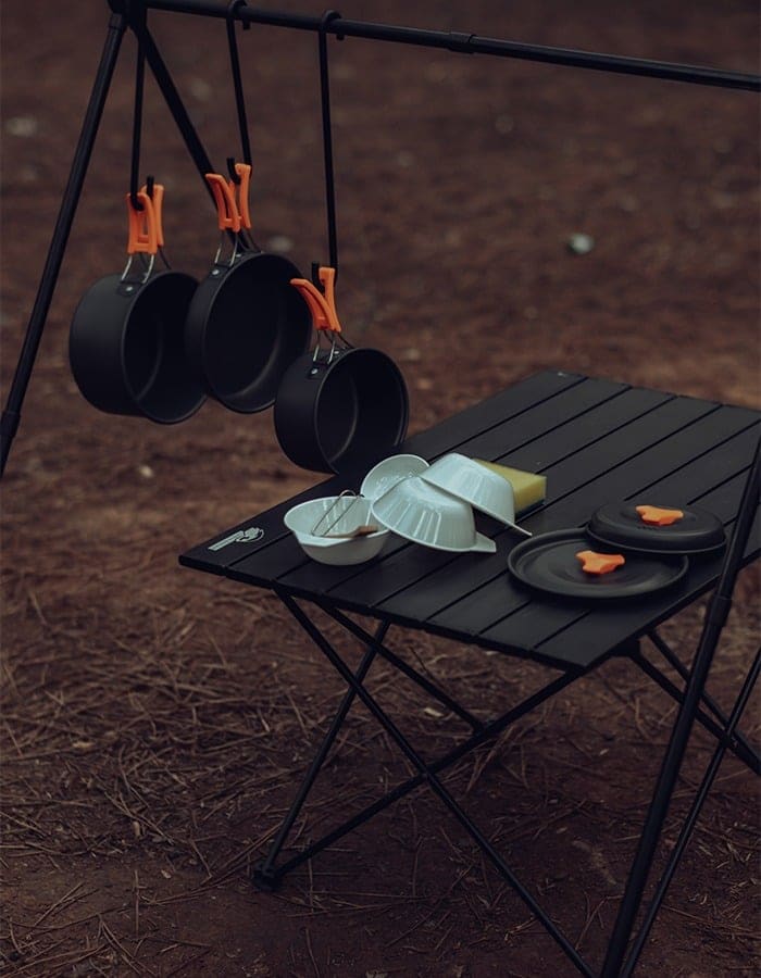 Pots and a pan with orange handles hanging from a rack above a camp table set with bowls, lids and a sponge