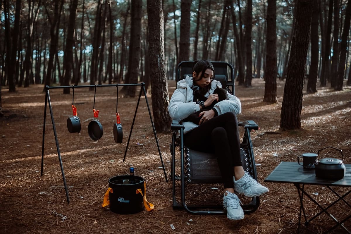 Woman holding a cat in a Heimdall zero gravity chair in a pine forest, a cook rack with pans beside her