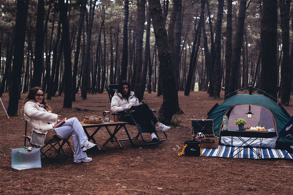 Two women relaxing at a pine forest camp with folding chairs, two tables, a tent and a striped picnic blanket