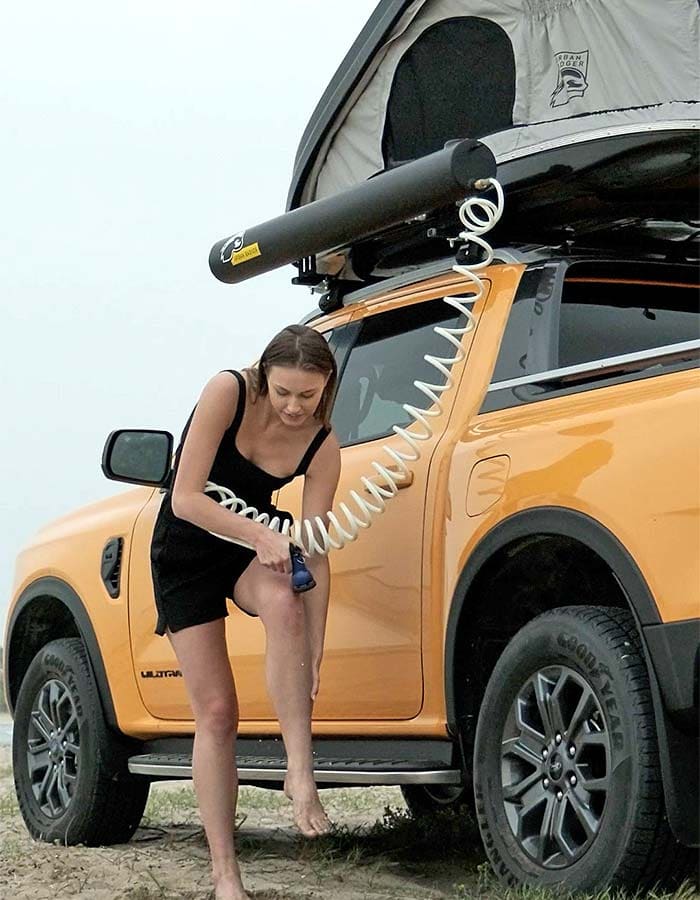 Woman rinsing her leg with the solar shower hose beside the orange pickup, the tank on the roof rack above