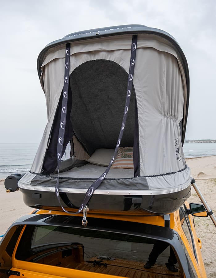 The open roof top tent on an orange pickup at the beach, seen from behind, with the mattress inside, the solar shower tank on the left and the ladder on the right