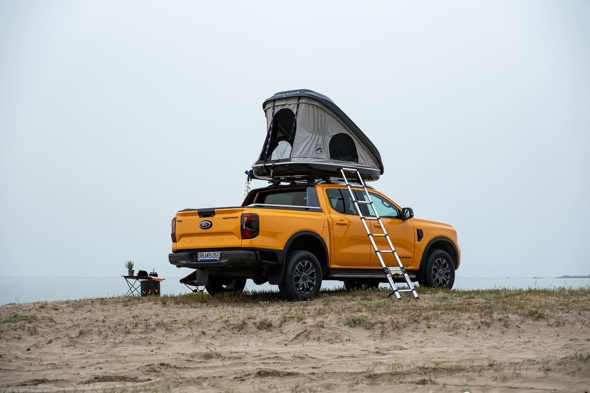 Telescopic ladder leaning on the open roof top tent of an orange pickup on a sandy rise by the sea