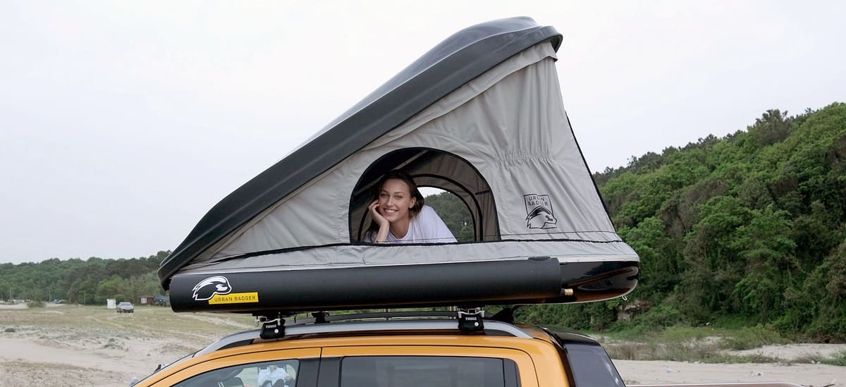 Woman smiling out of the side window of the roof top tent, wooded hills behind