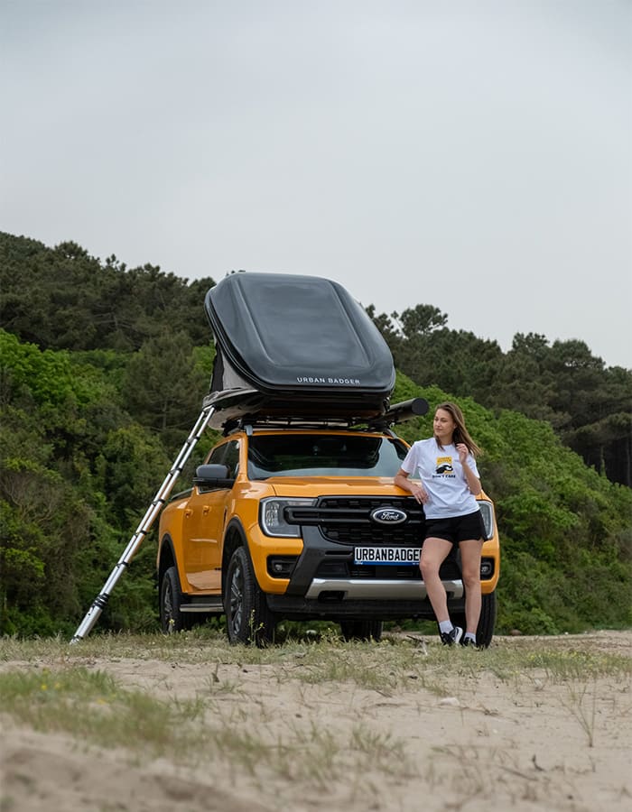 Woman leaning on the front of the pickup, the roof top tent on the roof rack and the ladder against it, wooded hill behind