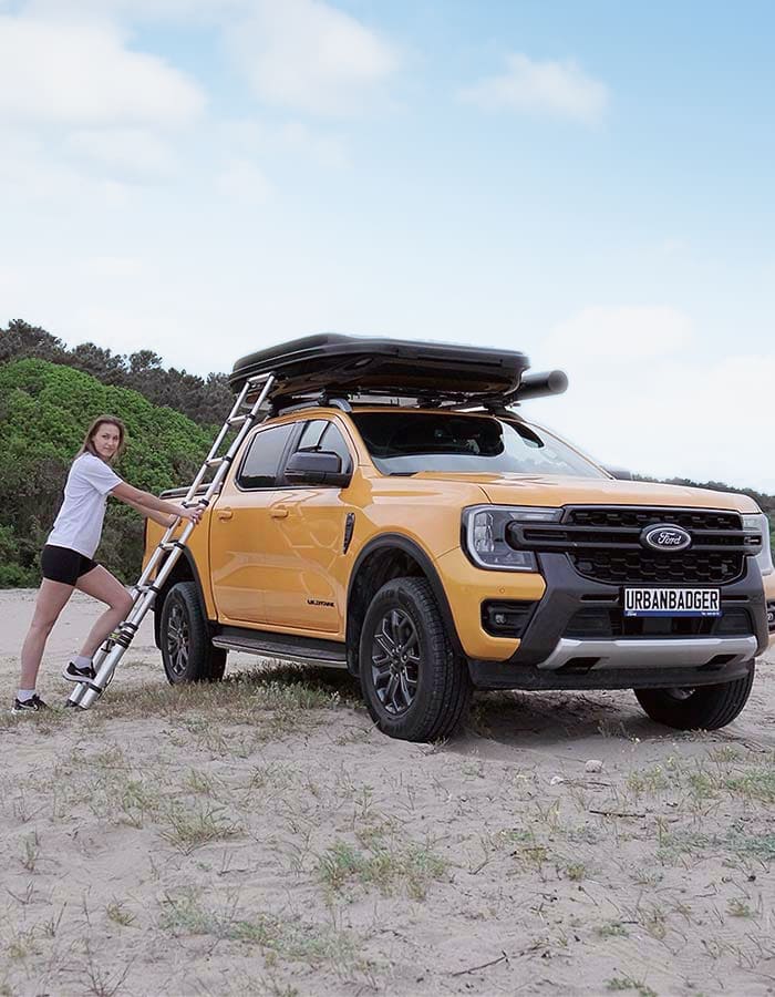Woman holding the extended telescopic ladder beside an orange pickup on the beach, the roof top tent on the roof rack