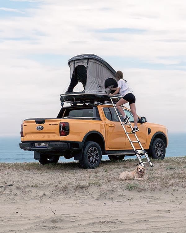 Woman climbing the telescopic ladder into a roof top tent on a pickup, a dog resting on the sand