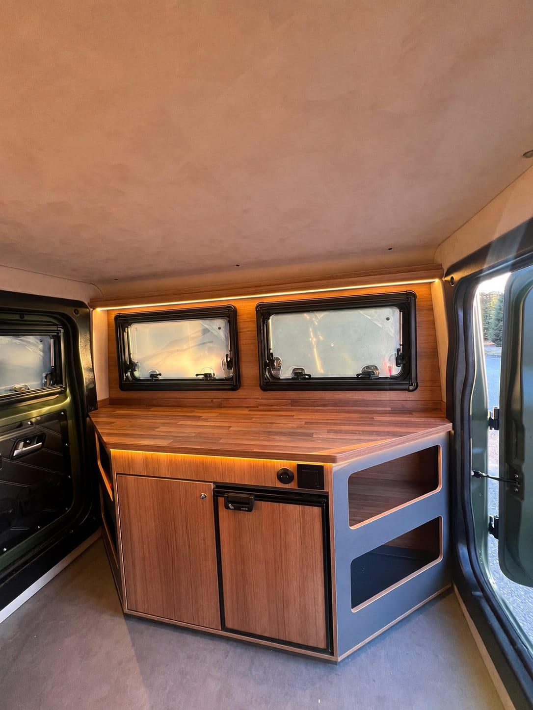 Wooden galley unit with cabinets and open shelves under two windows in the Urban Badger Towable Caravan