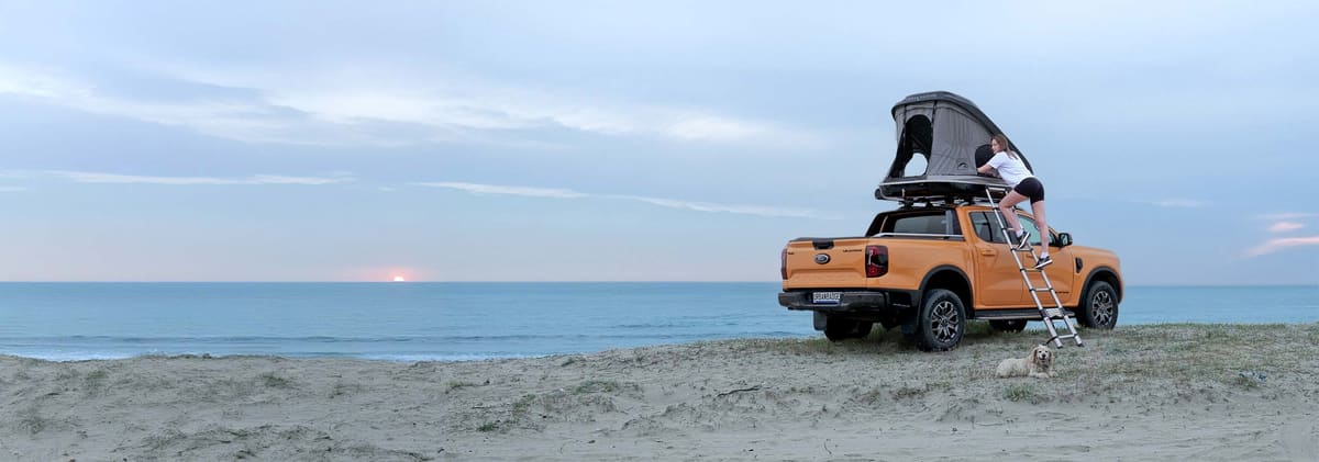 Woman climbing a telescopic ladder into an open roof top tent on a pickup at the beach at sunset, dog resting on the sand