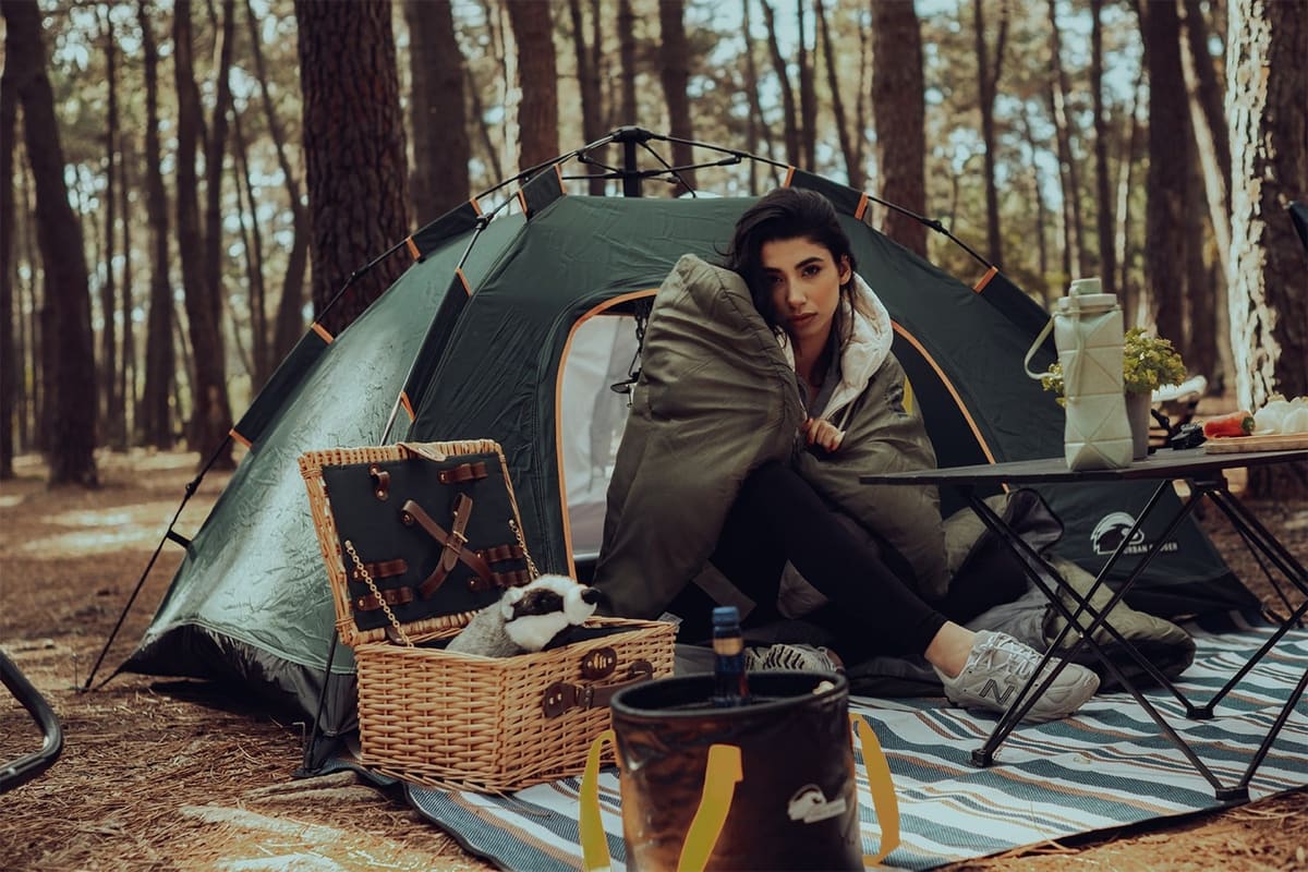 Woman wrapped in a Yule sleeping bag at the door of a Midgard tent in a pine forest, a picnic basket and a Wunjo blanket in front